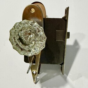 May include: A vintage door hardware set featuring a clear, faceted glass knob attached to a brass-toned metal backplate. The assembly includes a dark, rectangular iron mortise lock mechanism with a protruding latch and a small metal key inserted.