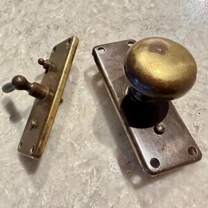 May include: A brass door knob set featuring a round knob mounted on a rectangular backplate with four screw holes, alongside a separate rectangular latch plate with a small lever handle and two screw holes, both showing a metallic finish.