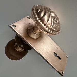 May include: A metallic door handle set featuring a rectangular backplate with a keyhole and a round, embossed knob. The front knob displays a decorative, scalloped pattern with a beaded border, while the rear knob is a simple, smooth, dark brown sphere.