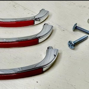 May include: Three curved drawer pulls with a polished chrome finish and a central red inlay. Each handle features two mounting points at the ends. Included are six silver-colored metal machine screws with flat heads for installation purposes.