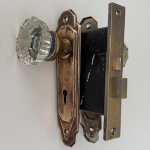 May include: A vintage mortise door lock set featuring a clear, faceted glass knob attached to a tarnished brass rectangular backplate with a keyhole. The internal mechanism is a black metal box with a brass latch and bolt on the side plate.