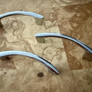 May include: Three polished chrome cabinet pulls with a curved, arching design. Each handle features a smooth, reflective metallic finish and two sturdy mounting points at the ends, providing a sleek and functional grip for drawers or doors.