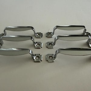 May include: A set of six polished chrome metal drawer pulls arranged in two columns of three. Each handle features a curved grip section and two flat mounting plates at the ends, each containing a single circular screw hole for installation.