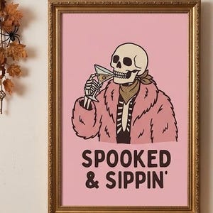 May include: A framed wall art print featuring a skeleton wearing a pink fur coat and a patterned neckerchief, holding a martini glass. The background is light pink with the black text SPOOKED & SIPPIN' printed at the bottom of the illustration.