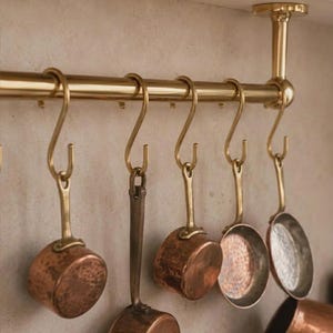 Peut inclure: Une tringle horizontale en laiton poli fixée au plafond avec cinq crochets en S assortis. Suspendues à ces crochets, plusieurs petites casseroles rondes en cuivre présentent des surfaces martelées et de courtes poignées métalliques.