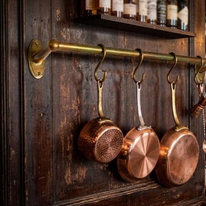 May include: A brass rail mounted on a dark wood wall with three S-shaped hooks holding copper pans. The pans have varying textures and finishes, including a hammered surface and smooth metallic bases. A shelf with glass bottles is visible above.