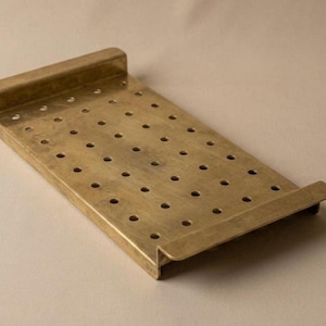 May include: A rectangular brass tray with a matte finish, featuring a grid of evenly spaced circular perforations across its surface and raised, flat-edged handles on both ends. The tray has a low-profile design with a consistent metallic tone.