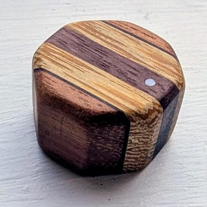 May include: A cylindrical wooden knob featuring a striped pattern of alternating light tan, medium brown, and dark purple wood grains. The top surface includes a small, circular white inlay dot positioned near the edge of the wood grain pattern.