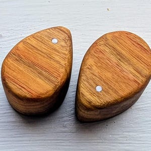 May include: A pair of teardrop-shaped wooden ear plugs with a smooth, polished finish. Each piece features a natural light brown wood grain pattern with subtle reddish-orange streaks and a single small, circular white inlay centered near the top.