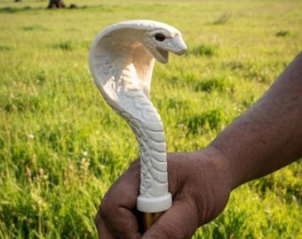 May include: A walking stick handle carved in the shape of a white cobra snake with a flared hood, detailed scale textures, and dark eyes. The handle is attached to a cylindrical shaft, featuring a smooth white finish and a metallic collar at the base.