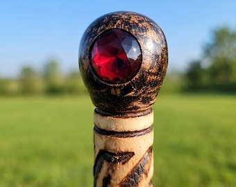 May include: A wooden staff featuring a spherical top with a dark, scorched finish and a faceted, deep red gemstone embedded in the center. The shaft below the sphere is lighter in color and displays a carved, spiraling pattern with dark accents.