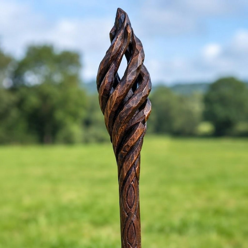 Puede incluir: Un bast&oacute;n de madera tallado a mano con un acabado marr&oacute;n oscuro y una secci&oacute;n superior texturizada en forma de llama en espiral. La superficie muestra marcas de cincel visibles y patrones de nudos tallados a lo largo del eje, creando un aspecto detallado.