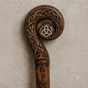 May include: A dark brown wooden staff featuring an intricate carved knotwork pattern on its curved top. A silver-colored triquetra symbol is embedded in the center of the spiral. The straight shaft below the curve displays carved runic characters.