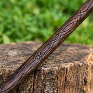 Puede incluir: Una varita de madera de color marr&oacute;n oscuro con un patr&oacute;n entrelazado tallado y repetitivo a lo largo de su longitud. La veta de la madera es visible en toda la superficie, mostrando un acabado pulido con surcos profundos que definen el dise&ntilde;o.