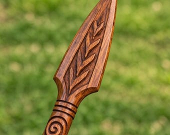 May include: A hand-carved wooden spearhead featuring a pointed tip and a symmetrical chevron pattern etched into the surface. The shaft below the head displays a circular spiral carving with horizontal bands, showcasing a rich brown wood grain.