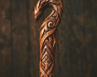 May include: A brown wooden walking stick featuring an intricate hand-carved design. The handle is shaped like a curved animal head with detailed textures, while the shaft displays geometric patterns and swirling motifs carved into the wood grain.
