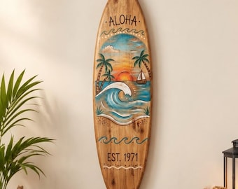 Peut inclure: Une décoration murale en forme de planche de surf en bois avec une peinture représentant un coucher de soleil, des palmiers, un voilier et une vague. Le mot ALOHA est inscrit en haut et le texte EST. 1971 figure en bas avec des lignes ondulées.
