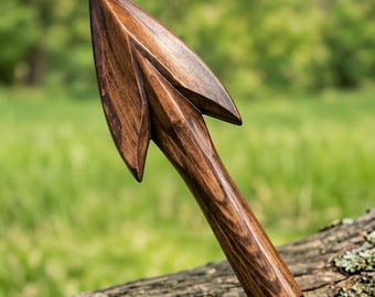 May include: A hand-carved wooden spearhead featuring a dark brown finish with visible wood grain patterns. The object has a sharp, pointed tip and two symmetrical barbs extending from the sides, tapering into a cylindrical shaft base.