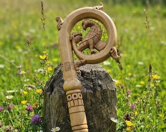 May include: A light brown wooden staff featuring a curved top with an intricate hand-carved design of a bird perched inside the loop. The shaft of the staff displays geometric patterns and horizontal bands carved into the wood surface.