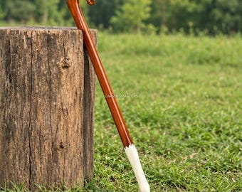 May include: A polished wooden walking stick featuring a carved horse head handle at the top. The shaft is a smooth, reddish-brown wood that transitions into a white, tapered ferrule at the base. The text CARVINGMASTERS is visible on the shaft.