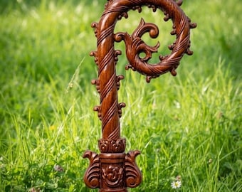 May include: A dark brown wooden crozier featuring a spiraled, curled top with intricate carved leaf-like details. The shaft is adorned with a decorative collar at the base of the crook, showcasing symmetrical floral carvings and a polished finish.