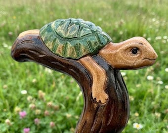 May include: A hand-carved wooden walking stick handle featuring a turtle with a green shell resting on a dark brown branch. The turtle has a light brown head and limbs, with a smooth finish and visible wood grain texture throughout the piece.