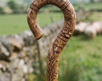 May include: A hand-carved wooden shepherd's crook featuring a curved handle with intricate leaf patterns and detailed relief carvings of small sheep. The natural brown wood grain is visible throughout the entire length of the staff.