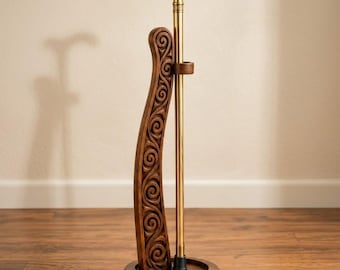 May include: A brass walking cane with an ornate handle rests in a dark wood stand featuring a carved spiral pattern. The stand has a circular base and a vertical support piece with a small ring to hold the cane upright. The text Carvingmasters is visible.