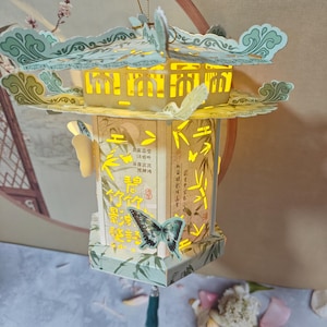 Puede incluir: Una linterna de papel hexagonal con techo escalonado, iluminada desde el interior. La estructura presenta patrones de bambú verde claro, formas de mariposas recortadas y caracteres chinos impresos en los paneles. Una mariposa está fijada al frente.