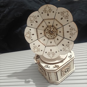 Puede incluir: Un modelo de gramófono de madera clara con una bocina circular grande de varios paneles que presenta patrones decorativos cortados con láser. La base es una caja hexagonal con intrincados diseños recortados en los lados y una manivela.