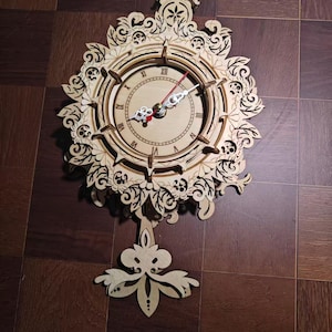 Puede incluir: Un reloj de pared de madera clara con intrincados patrones florales y de volutas cortados con láser. La esfera muestra números romanos, con un segundero rojo y manecillas de hora y minuto plateadas. Un péndulo decorativo cuelga bajo la esfera.