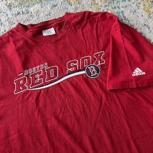 Peut inclure: Un t-shirt rouge à manches courtes avec le texte BOSTON RED SOX en blanc et contours bleu marine. Un logo circulaire avec un B rouge se trouve à droite du texte. Un logo Adidas est imprimé en blanc sur la manche gauche du vêtement.