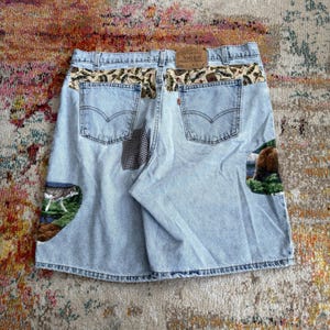 Pode incluir: Calções de ganga azul-claro com uma etiqueta de couro bege na cintura, dois bolsos traseiros e painéis de tecido decorativos. Os painéis exibem um padrão de peixes, um lobo numa paisagem florestal e um urso castanho num ambiente arborizado.