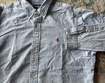 May include: A light blue long-sleeve button-down shirt featuring a button-down collar and a red embroidered logo on the chest. The shirt has a central placket with light-colored buttons and a label at the neck that reads Ralph Lauren.