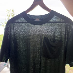 May include: A dark green short-sleeved t-shirt with a crew neckline and a single chest pocket on the left side. The fabric has a semi-sheer, lightweight texture. The shirt is displayed on a wooden hanger with the text LEVY'S APPAREL STORE FOR MEN.