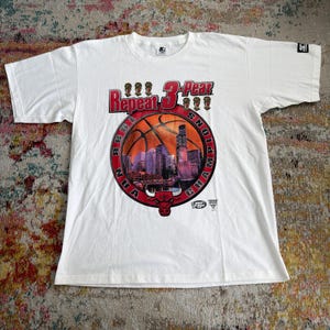 May include: A white short-sleeve t-shirt featuring a graphic of the Chicago skyline inside a basketball, with the text Repeat 3-Peat and 1998 NBA Champions. The design includes a red bull logo, trophy icons, and a Starter brand logo on the sleeve.
