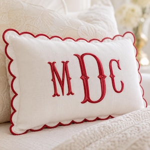 May include: A rectangular off-white throw pillow featuring a scalloped edge trimmed in red thread. The center of the pillow displays a large, embroidered monogram in red thread with the letters M, D, and C in a decorative serif font style.