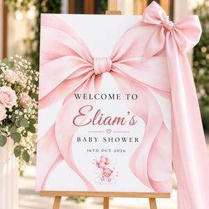 May include: A white rectangular baby shower welcome sign featuring a large, soft pink painted bow graphic. The text reads: WELCOME TO Eliam's BABY SHOWER 16TH OCT 2026. Below the text is a small illustration of a teddy bear holding pink balloons.