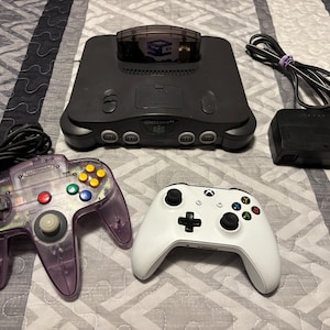 May include: A black Nintendo 64 game console with a game cartridge inserted, a translucent purple wired controller, a white wireless controller, and a black power adapter with a cord. The console features the Nintendo 64 logo on the front panel.