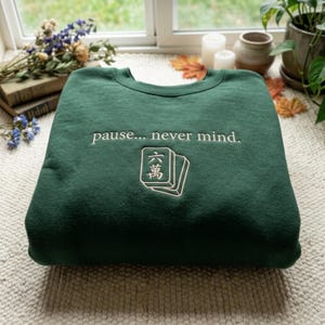 Könnte beinhalten: Ein dunkelgrünes Sweatshirt mit Rundhalsausschnitt und dem gestickten Text "pause... never mind." in hellem Cremeweiß. Unter dem Text befindet sich eine Stickerei von zwei Mahjong-Steinen, wobei der obere Stein das chinesische Schriftzeichen für sechs zeigt.