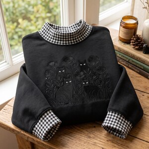 May include: A black long-sleeve sweatshirt featuring a mock neck collar and sleeve cuffs in a black and white gingham pattern. The front of the garment displays an embroidered design of three black cats sitting among flowers and tall grass.