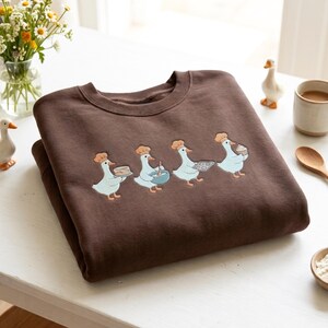 May include: A dark brown crewneck sweatshirt featuring an embroidered graphic of four white geese wearing tan chef hats. The geese are depicted in a line, each performing a baking task: holding a cake, mixing a bowl, carrying a tray, and holding a cupcake.