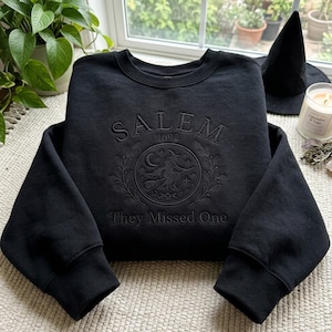 Könnte beinhalten: Ein schwarzes Sweatshirt mit Rundhalsausschnitt und tonaler Stickerei auf der Brust. Das Design zeigt den Schriftzug SALEM 1692, ein kreisförmiges Emblem mit einer Hexe auf einem Besen, Sternen, einer Mondsichel und den Text They Missed One.