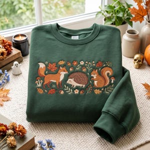 May include: A dark green crewneck sweatshirt featuring an embroidered design of a fox, a hedgehog, and a squirrel surrounded by autumn leaves, acorns, and small mushrooms. The embroidery is centred on the chest of the long-sleeved garment.