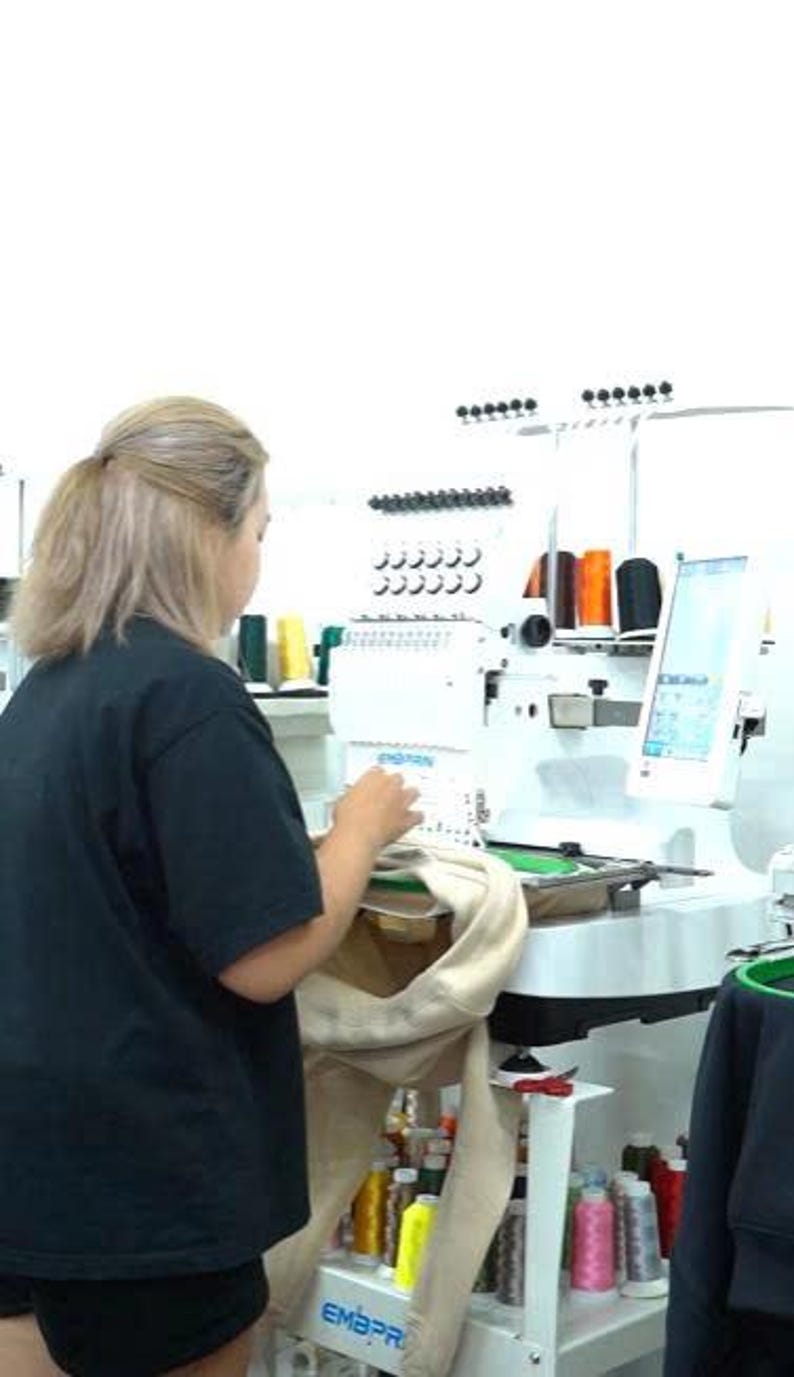 May include: A white industrial embroidery machine with a digital screen and multiple thread spools in various colors. The machine is mounted on a white metal stand with shelves holding additional spools of thread and a beige garment in the hoop.