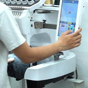 May include: A white industrial embroidery machine featuring a digital touchscreen interface, multiple thread spools in various colors, and a needle bar assembly. The machine is set up with a dark fabric item positioned for embroidery work.