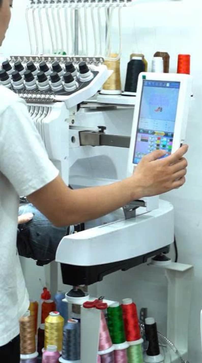 May include: A white industrial embroidery machine featuring a digital touchscreen interface, multiple thread spools in various colors, and a needle bar assembly. The machine is set up with a dark fabric item positioned for embroidery work.