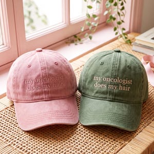 Peut inclure: Deux casquettes de baseball, une rose poudré et une vert olive, arborant toutes deux le texte brodé « my oncologist does my hair » en fil clair. Chaque casquette présente une calotte structurée, une visière courbée et des œillets cousus.