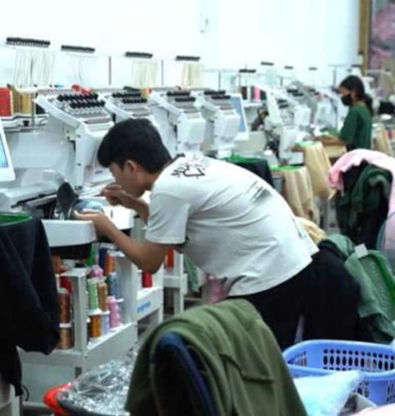May include: A person in a white t-shirt operates a white industrial embroidery machine in a workshop. The machine features multiple spools of colorful thread and needles. Other similar machines are visible in the background of the room.