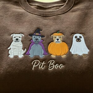 May include: A brown sweatshirt featuring embroidered designs of four dogs in Halloween costumes: a mummy, a witch with a purple cape, a pumpkin, and a ghost. Below the dogs, the text Pit Boo is embroidered in a white cursive script font.
